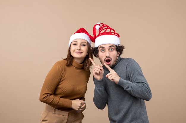 Concepto de fiesta y fiesta de vacaciones - feliz encantadora pareja joven sorprendida vistiendo unidos unos a otros sombreros de santa claus en imágenes grises