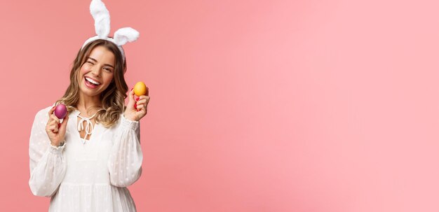 Concepto de fiesta y primavera de vacaciones Alegre mujer rubia guapa celebrando el día de Pascua en orejas de conejo sosteniendo dos huevos pintados y una cámara de guiño sonriendo felizmente fondo rosa