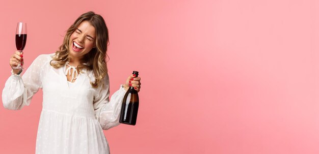 Concepto de fiesta y primavera de vacaciones Retrato de una chica rubia guapa emocionada y emotiva bailando y celebrando divertirse diciendo sí cantando con los ojos cerrados sostenga una copa de vino y una botella