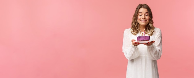 Concepto de fiesta y primavera de vacaciones Retrato de una soñadora feliz cumpleaños que siente emoción y alegría celebrando el cumpleaños mordiéndose el labio y sonriendo mientras pide un deseo soplando una vela en el pastel