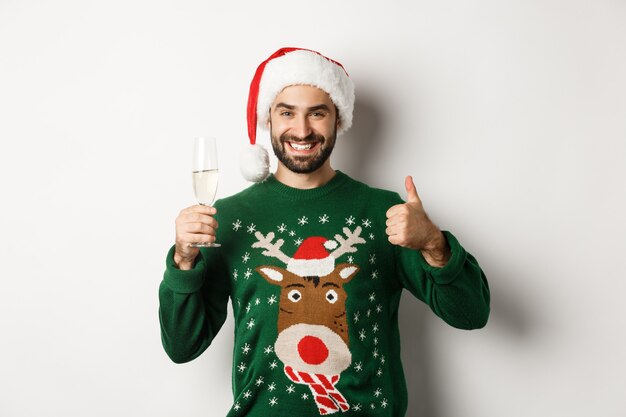 Concepto de fiesta y vacaciones de Navidad. Chico satisfecho con gorro de Papá Noel y suéter mostrando los pulgares para arriba y bebiendo una copa de champán, de pie sobre fondo blanco.