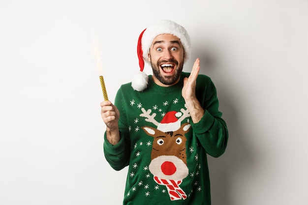 Concepto de fiesta y vacaciones de Navidad. Feliz joven celebrando el año nuevo de Navidad, sosteniendo la bengala y mirando emocionado, de pie sobre fondo blanco.