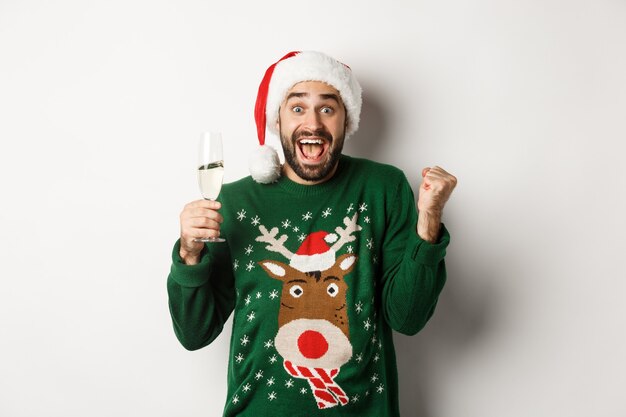 Concepto de fiesta y vacaciones de Navidad. Hombre emocionado con gorro de Papá Noel celebrando el Año Nuevo, bebiendo champán y regocijándose, de pie sobre fondo blanco.