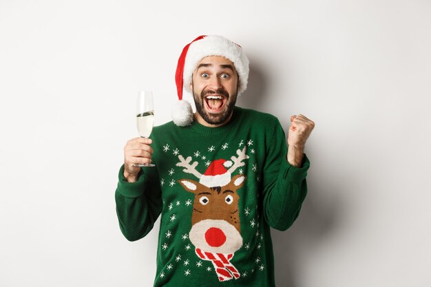 Concepto de fiesta y vacaciones de Navidad. Hombre emocionado con sombrero de Santa celebrando el año nuevo, bebiendo champán y regocijándose, de pie sobre fondo blanco.
