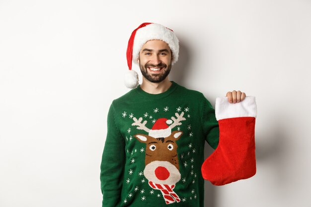 Concepto de fiesta y vacaciones de Navidad. Hombre feliz con sombrero de Santa trayendo regalos en calcetín de Navidad y sonriendo, de pie sobre fondo blanco.
