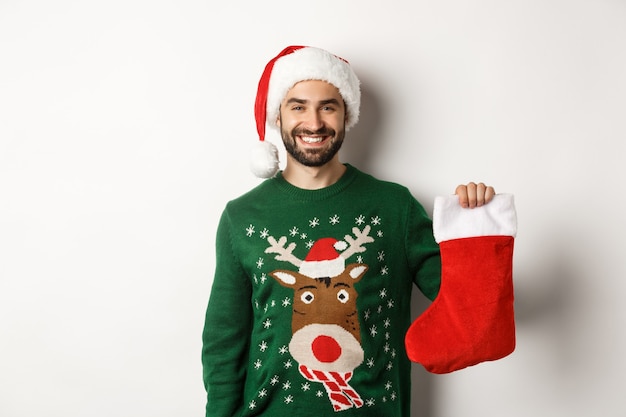 Concepto de fiesta y vacaciones de Navidad. Hombre feliz con sombrero de Santa trayendo regalos en calcetín de Navidad y sonriendo, de pie sobre fondo blanco.