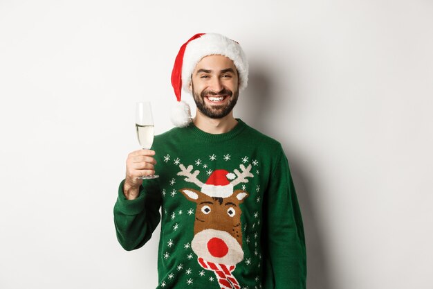 Concepto de fiesta y vacaciones de Navidad. Hombre guapo con barba con gorro de Papá Noel y suéter divertido, bebiendo champán y celebrando el año nuevo, fondo blanco.