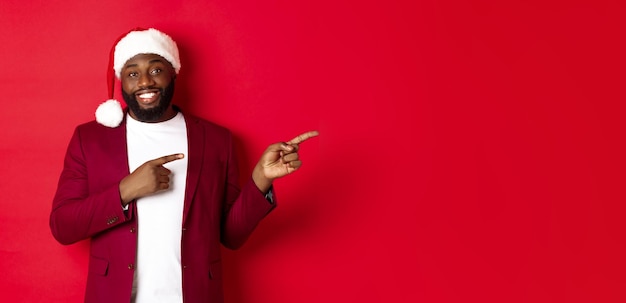 Foto gratuita concepto de fiesta y vacaciones de navidad hombre negro sonriente con barba y sombrero de santa apuntando con el dedo aparejo