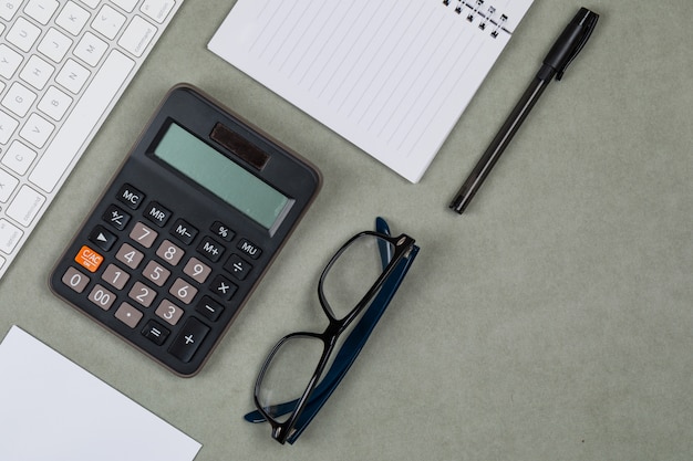 Concepto financiero con cuaderno, papel, bolígrafo, calculadora, teclado, gafas sobre fondo gris plano lay.