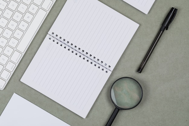 Concepto financiero con cuadernos, papel, bolígrafo, lupa, teclado sobre fondo gris endecha plana.