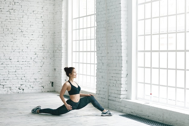 Foto gratuita concepto de fitness, deportes, energía, salud y actividad. imagen de una alegre chica atlética en elegantes zapatillas, top y leggings haciendo ejercicio en el interior, estirando las piernas frente a una ventana grande