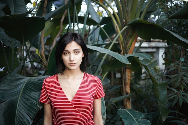 Concepto de frescura, belleza y cosmética natural. Toma al aire libre de modelo de chica impresionante con cabello bob oscuro posando entre plantas exóticas, promocionando crema facial orgánica u otro producto cosmético para el cuidado de la piel