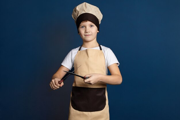 Concepto de gastronomía, cocina, catering e industria alimentaria. Foto de estudio de guapo lindo niño de 10 años vestido con cuchillo de afilar uniforme de chef con otro. Niño varón afilado cuchillos de cocina