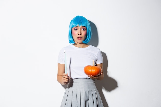 Concepto de halloween. Imagen de una niña asiática asustada con peluca azul que parece nerviosa y asustada, sosteniendo una vela y una calabaza.