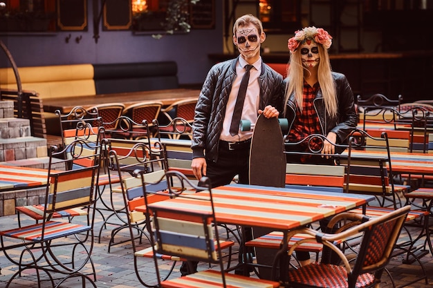 Concepto de Halloween y Muertos. Joven pareja atractiva con muertos vivientes posando con longboard cerca de un restaurante mexicano al aire libre.