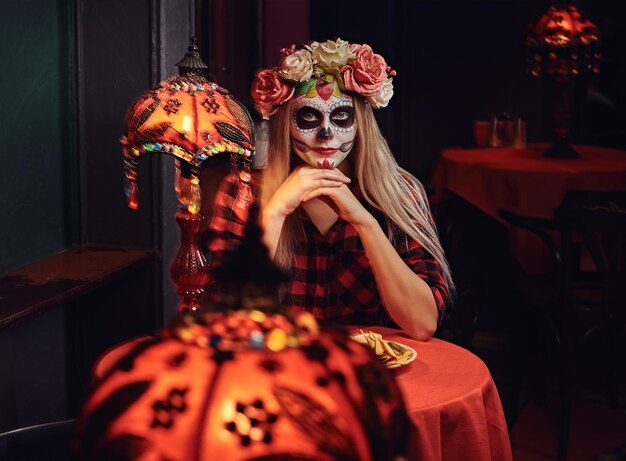 Concepto de Halloween y Muertos. Joven rubia con maquillaje no muerto en corona de flores comiendo nachos en un restaurante mexicano.
