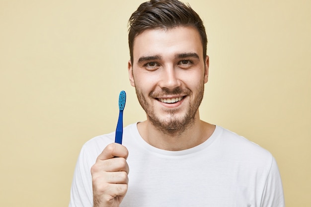 Concepto de higiene dental y área bucal saludable. Retrato de atractivo joven feliz haciendo rutina matutina posando aislado con cepillo de dientes, yendo a limpiar los dientes antes de dormir, mirando con sonrisa