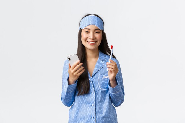 Concepto de higiene, estilo de vida y personas en el hogar. Sonriente linda chica asiática en pijama azul y máscara para dormir, cepillándose los dientes antes de acostarse y usando el teléfono inteligente, mostrando dientes blancos, fondo blanco.