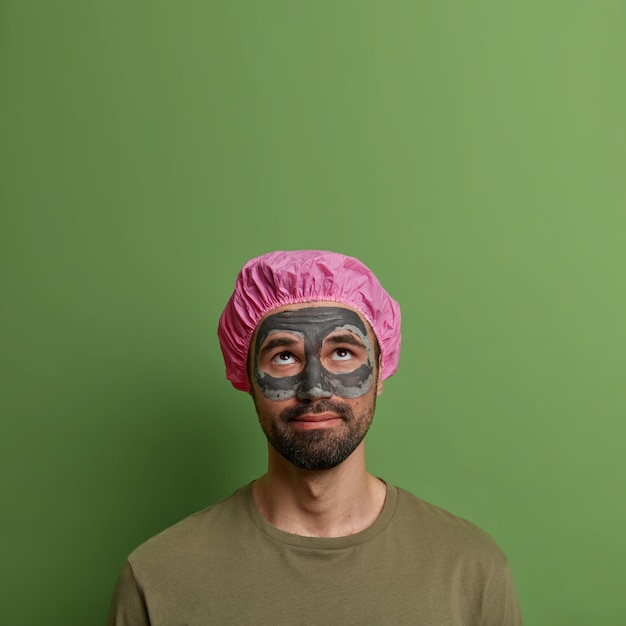 Concepto de hombres, cosmetología, higiene y belleza. Hombre serio y atento concentrado arriba, aplica mascarilla de arcilla en la cara para rejuvenecer, tiene barba densa, usa gorro de ducha, mira el espacio vacío hacia arriba