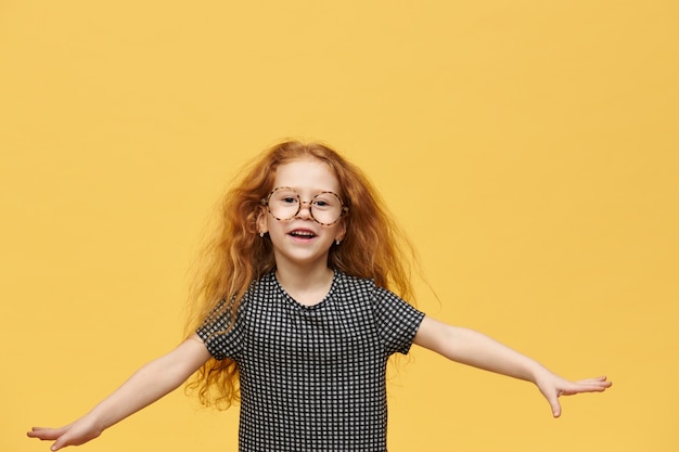 Concepto de infancia, diversión y alegría. Adorable niña emocional con voluminoso cabello jengibre exclamando con entusiasmo, saltando, manteniendo los brazos abiertos posando en la pared amarilla en blanco con copyspace para su texto