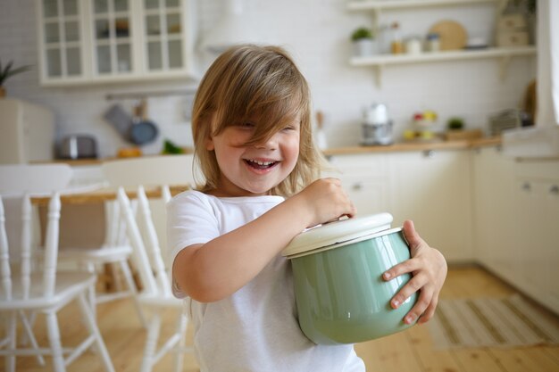 Concepto de infancia e inocencia. Hermosa niña adorable con sonrisa encantadora, jugando con cazuela de madres. Linda niña sosteniendo una cacerola, yendo a cocinar sopa para la cena, sonriendo felizmente