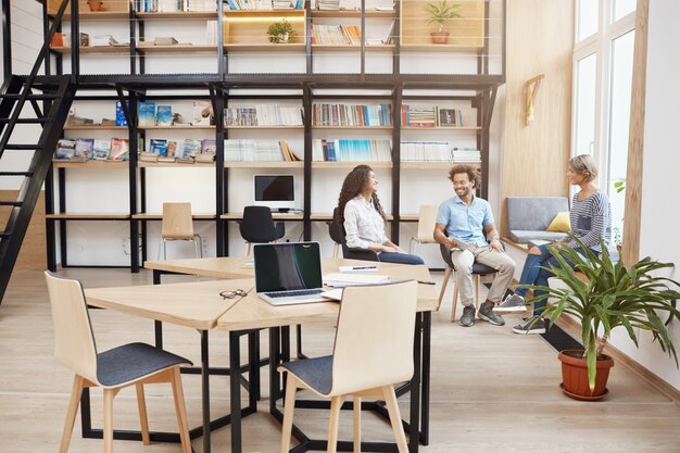 Concepto de inicio, negocios, trabajo en equipo. Grupo de jóvenes de perspectiva en una reunión en una gran biblioteca moderna hablando sobre las ganancias del último proyecto, mirando documentos, sonriendo y teniendo un tiempo productivo