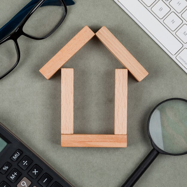 Concepto inmobiliario con casa hecha de bloques de madera, gafas, lupa, teclados en primer plano de fondo gris.