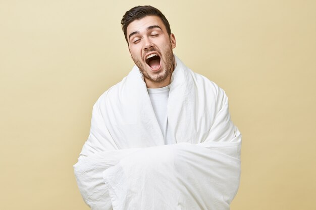 Concepto de insomnio y privación del sueño. Retrato de hombre joven con párpados adormilados bostezando con la boca abierta posando aislado envuelto en una manta blanca, sintiéndose somnoliento debido al despertar temprano