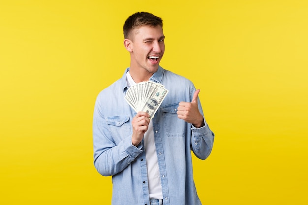 Concepto de inversión, compras y finanzas. Hombre rubio guapo descarado que muestra el pulgar hacia arriba y guiña un ojo, sonriendo para animar a probar la lotería o el casino, de pie sobre un fondo amarillo.