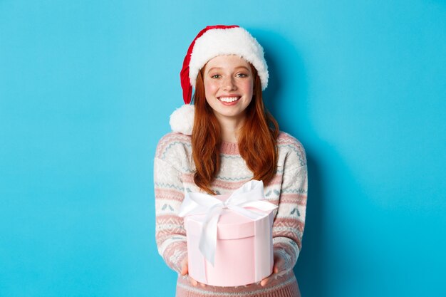 Concepto de invierno y celebración. Hermosa chica pelirroja con sombrero de santa deseando feliz Navidad, dando regalos y sonriendo, de pie sobre fondo azul.