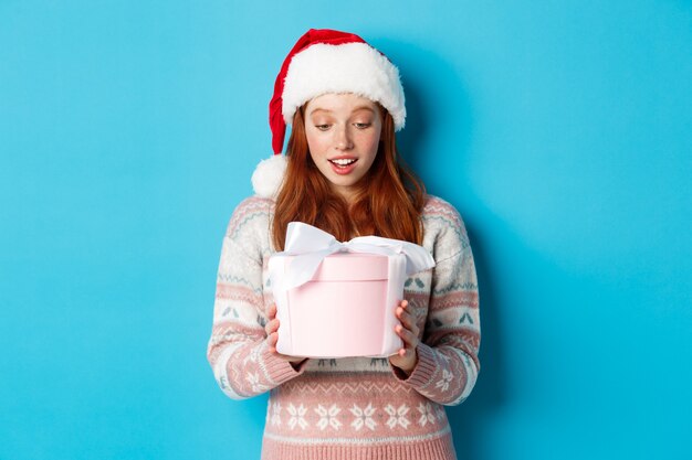 Concepto de invierno y Nochebuena. Chica pelirroja conmovida y halagada mirando la caja con regalo de Navidad, sonriendo asombrada, de pie con gorro de Papá Noel contra el fondo azul.