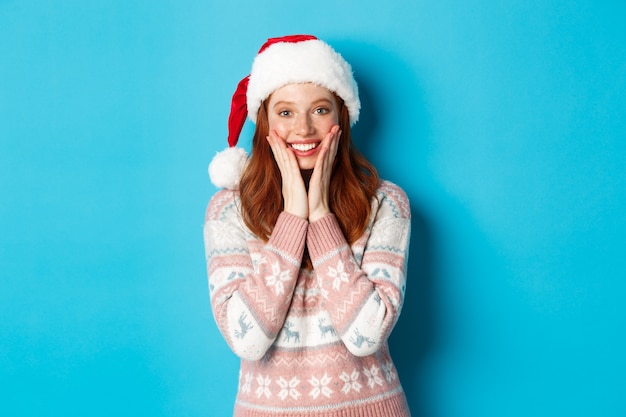 Concepto de invierno y Nochebuena. Chica pelirroja feliz celebrando la Navidad, mirando a la cámara asombrada, sonriendo y tocando las mejillas, de pie sobre fondo azul.