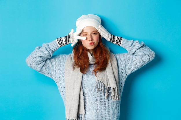 Concepto de invierno y vacaciones. Feliz chica pelirroja con gorro, bufanda y guantes mostrando el signo de la paz y los labios fruncidos descarados, mirando a la cámara, vistiendo un suéter, posando sobre fondo azul