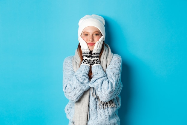 Concepto de invierno y vacaciones. Linda chica en gorro blanco, suéter y guantes, apretando las mejillas y sonriendo complacido, calentamiento después del frío al aire libre, de pie sobre fondo azul