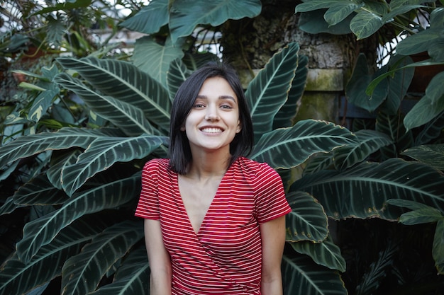 Concepto de juventud, belleza, naturaleza y ecología. Retrato de feliz elegante joven ecologista europea trabajando al aire libre, posando contra las plantas tropicales verdes, con expresión alegre en su rostro