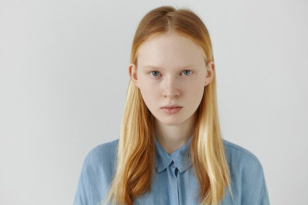 Concepto de juventud y belleza. Retrato de la hermosa adolescente caucásica con su largo cabello rubio hacia atrás mirando con expresión facial grave. Linda mujer joven con pecas posando en el interior