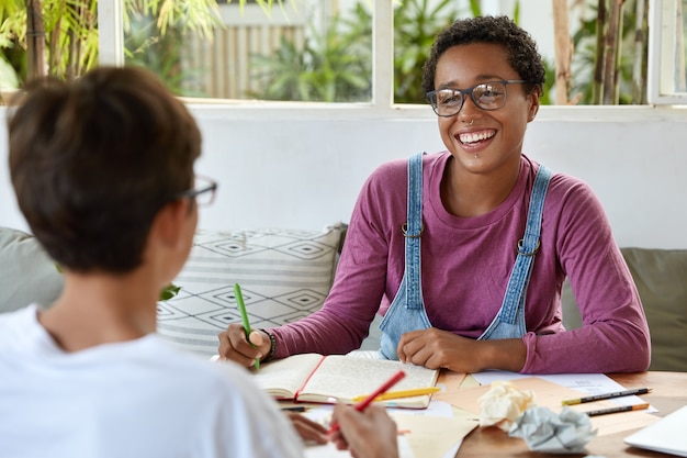 Concepto de juventud y coworking. Dama de piel oscura y sonriente complacida con anteojos, usa piercings, colabora con su compañera de grupo, hace tareas comunes, prepara tareas en casa, discute temas educativos.