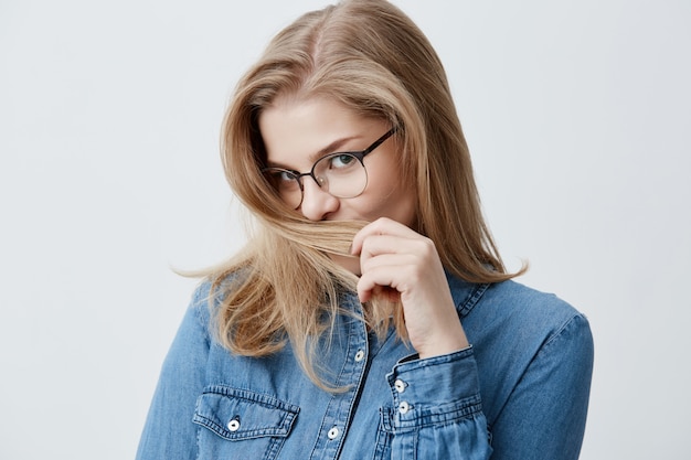 Concepto de juventud y felicidad. Hermosa adolescente caucásica en camisa vaquera, mirando a la cámara con atractivo, jugando con el pelo rubio. Mujer joven con una piel sana perfecta en gafas con estilo