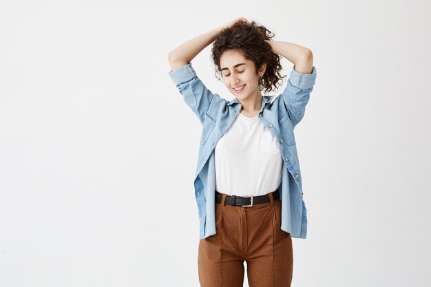 Concepto de juventud y felicidad. Morena adolescente en camisa vaquera y pantalón marrón, posando contra la pared blanca con los ojos cerrados, jugando con su largo cabello ondulado. La mujer joven sonríe agradablemente