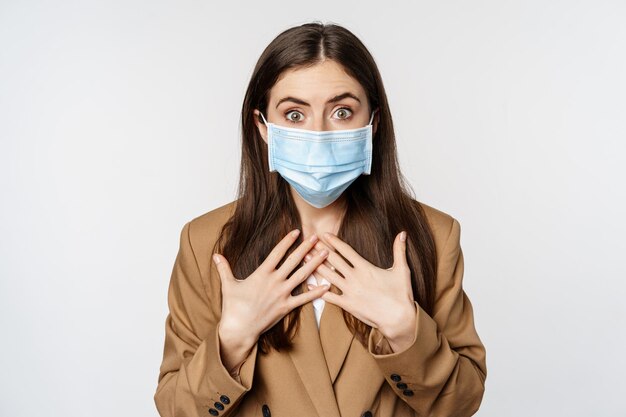 Concepto de lugar de trabajo y pandemia. Mujer de negocios conmocionada con mascarilla médica, jadeando, sorprendida y preocupada por la cámara, de pie sobre fondo blanco.