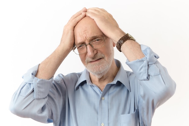 Concepto de malas noticias, estrés y personas. Foto de estudio de frustrado hombre caucásico de sesenta años con ropa formal y gafas sosteniendo la mano en la cabeza, habiendo subrayado la mirada triste debido a problemas