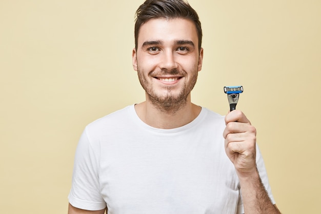 Concepto de masculinidad, belleza y cuidado de la piel. Retrato de hombre joven guapo con cerdas con amplia sonrisa sosteniendo el palo de afeitar, yendo a afeitarse la barba por la mañana antes del trabajo