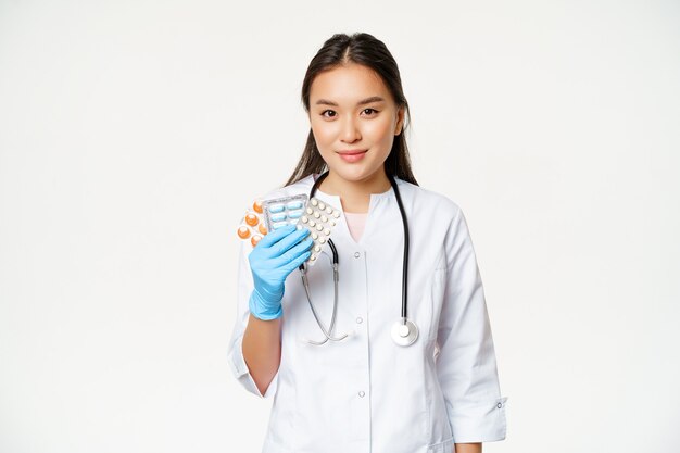 Concepto médico y sanitario. Doctora asiática mostrando píldoras, vitaminas en guantes de goma estériles, de pie en uniforme sobre fondo blanco.