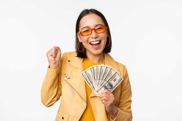 Concepto de microcrédito y dinero Elegante mujer joven asiática en gafas de sol riendo feliz sosteniendo dólares en efectivo de pie sobre fondo blanco.