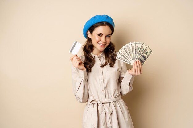 Concepto de microcrédito y dinero. Mujer joven y elegante que muestra tarjeta de crédito y dólares en efectivo, sonriendo feliz y satisfecha, de pie sobre un fondo beige.