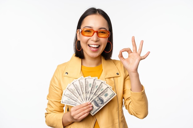 Concepto de microcrédito y préstamos Feliz chica coreana con estilo que muestra el signo de ok y dinero en efectivo de pie en ropa de moda sobre fondo blanco.