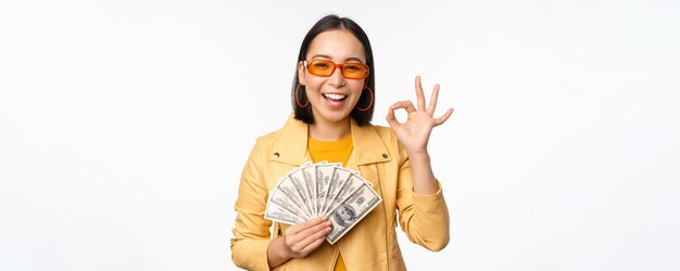 Concepto de microcrédito y préstamos Feliz chica coreana con estilo que muestra el signo de ok y dinero en efectivo de pie en ropa de moda sobre fondo blanco.