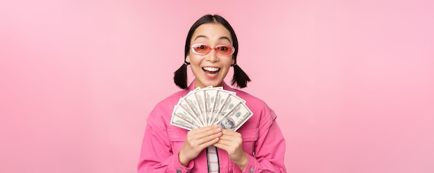 Concepto de microcrédito y préstamos rápidos Chica coreana elegante y emocionada que muestra dinero en efectivo y se ve feliz de pie con gafas de sol sobre fondo rosa