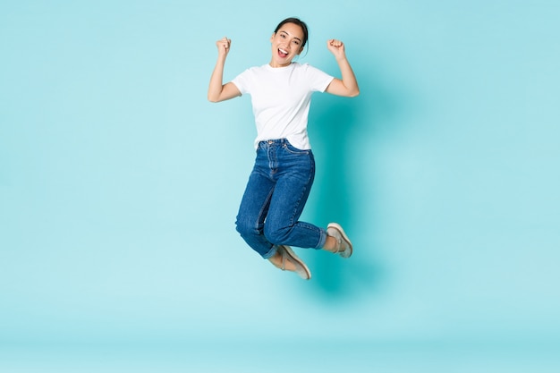 Concepto de moda, belleza y estilo de vida. Alegre, triunfante, atractiva chica asiática saltando de felicidad y alegría, ganando la competencia, celebrando la victoria sobre la pared azul claro