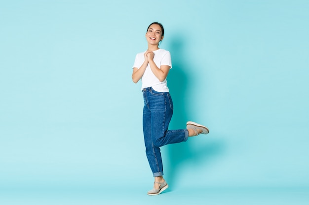 Concepto de moda, belleza y estilo de vida. Hermosa chica asiática romántica y soñadora en traje casual con aspecto encantador, unir las manos juntas posando sobre la pared azul claro.